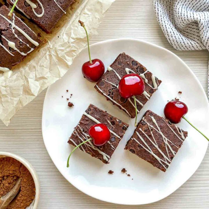 Chocolate beer brownies drizzled with white chocolate and topped with fresh cherries, served on a white plate with parchment-lined brownies in the background