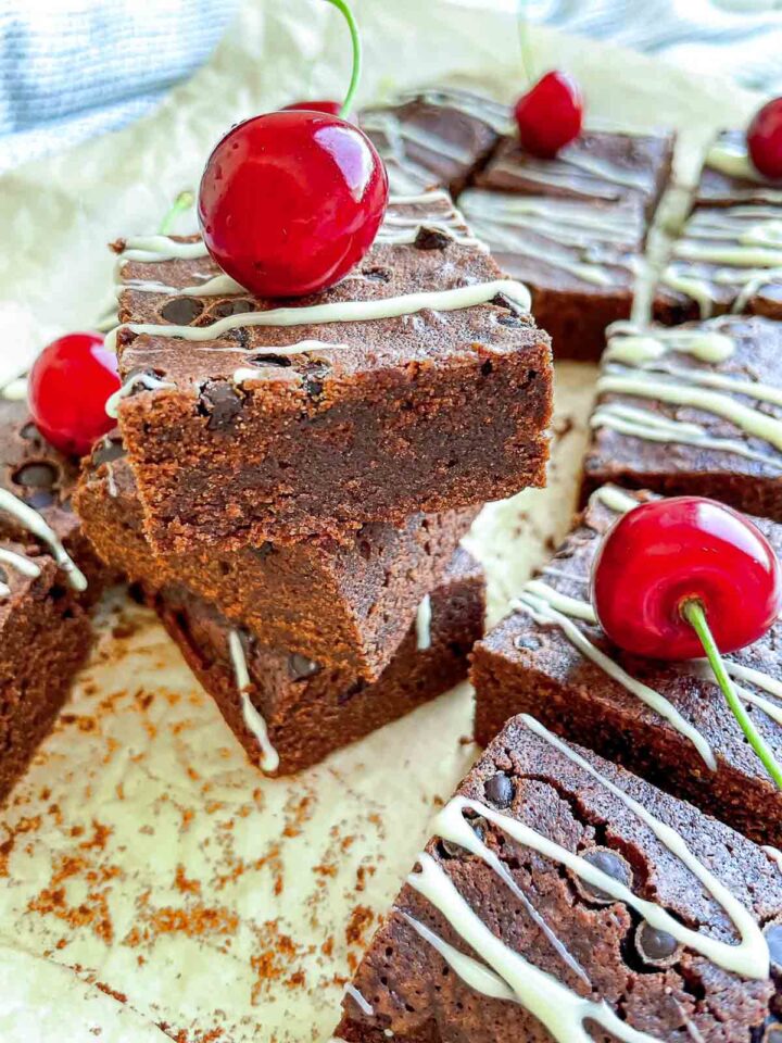 Stacked chocolate beer brownies drizzled with white chocolate and topped with glossy red cherries on parchment paper