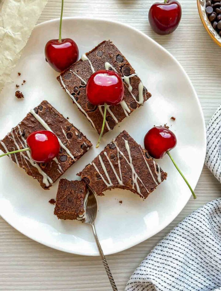 Chocolate beer brownies drizzled with white chocolate and topped with fresh cherries, served on a white plate with a piece cut using a spoon.