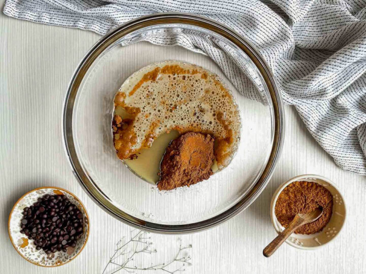 Glass mixing bowl with beer, melted butter, and cocoa powder being combined on a light surface, with chocolate chips and cocoa nearby