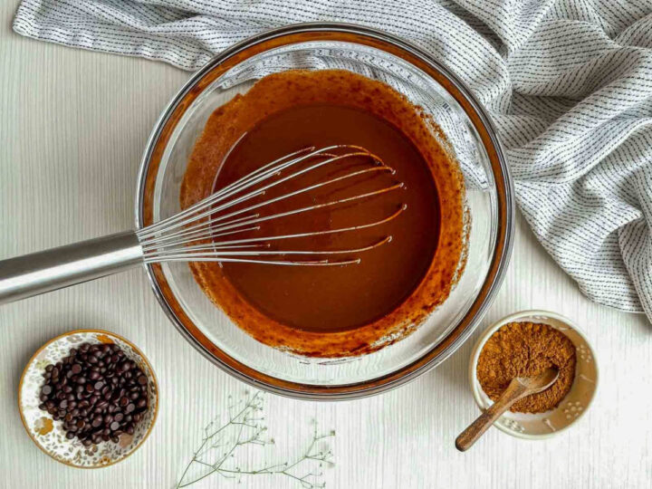 Chocolate brownie batter being whisked smooth in a clear glass bowl, with cocoa powder and chocolate chips nearby on a light kitchen surface