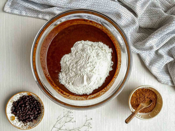 All-purpose flour added to a bowl of chocolate brownie batter, with chocolate chips and cocoa powder nearby on a light kitchen surface
