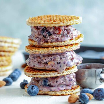 Blueberry ice cream waffle sandwiches stacked with rainbow sprinkles, fresh blueberries, and nuts on a light surface.