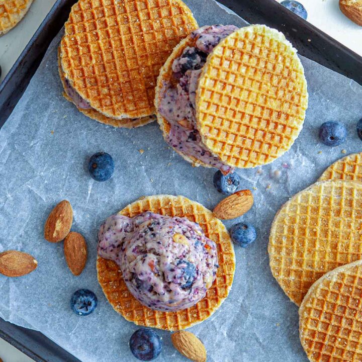 Scoops of blueberry ice cream placed between crisp waffle cookies on a baking tray, with fresh blueberries and almonds scattered around