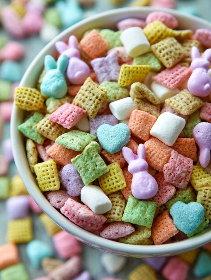Close-up of bunny bait snack mix in a white bowl with pastel cereal squares, mini marshmallows, bunny-shaped marshmallows, and colorful Easter candy on a white marble surface, tightly cropped to show texture and Easter colors.