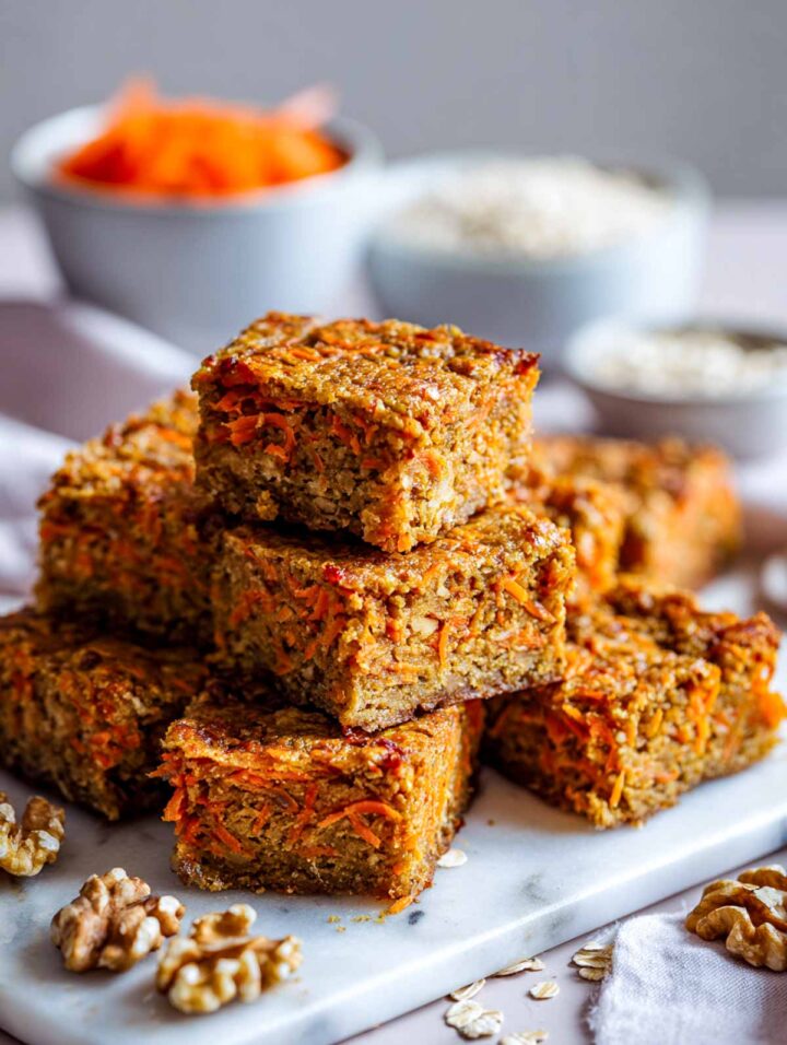 Stacked carrot cake energy squares with visible shredded carrot and oats, arranged on a white marble plate with grated carrots, oats, and walnuts nearby.