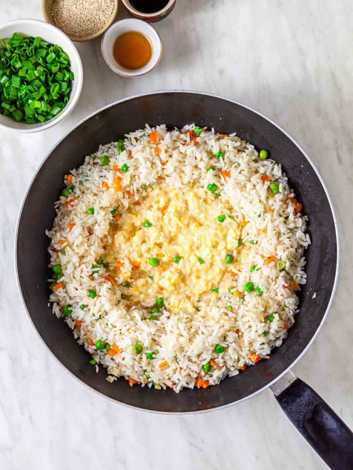 Scrambled eggs being mixed into fried rice with peas and carrots in a skillet.
