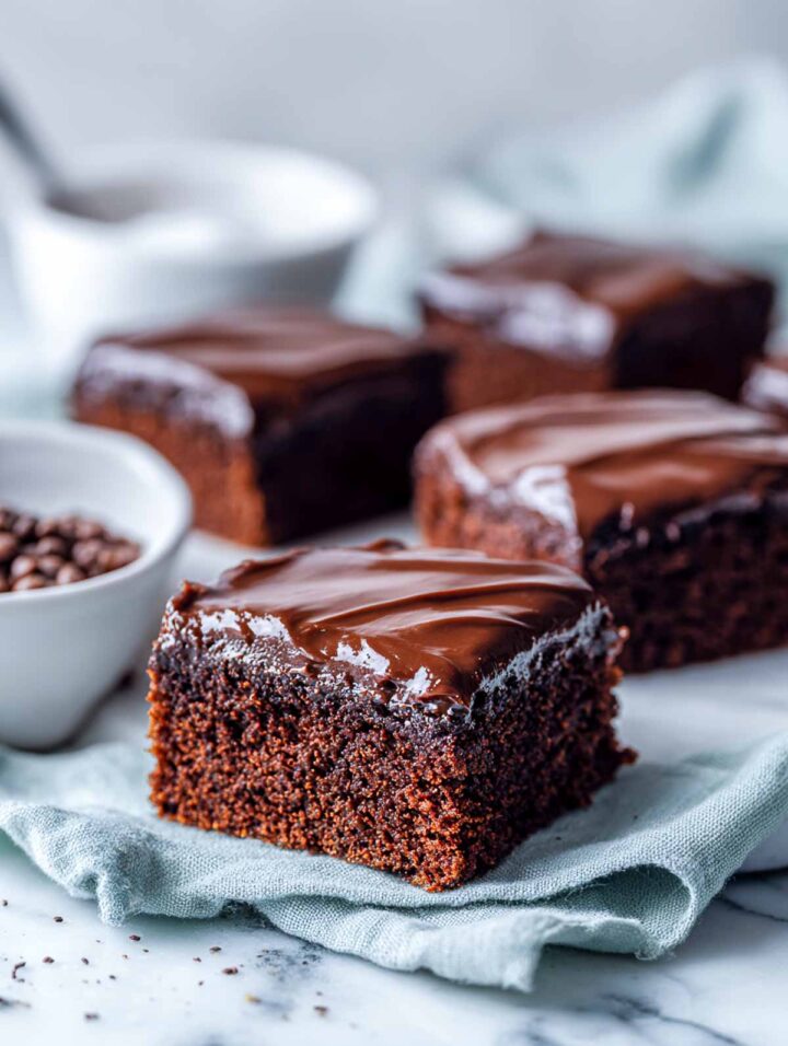 Chocolate chickpea snack cake cut into neat squares and topped with a smooth fudge frosting, arranged on a white marble surface with a soft sage napkin. Small white bowls with cocoa powder and cooked chickpeas sit nearby for a clean, bright, bakery-style presentation.