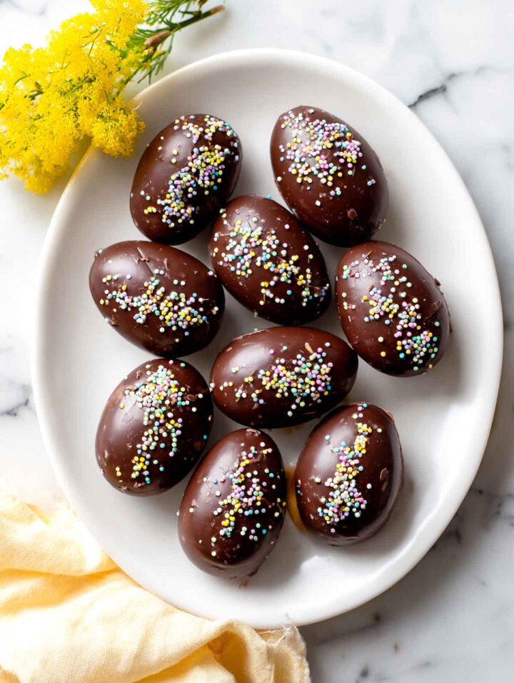 Top-down view of chocolate sunflower butter eggs arranged on a white plate, topped with spring sprinkles, with one egg cut open to show a sunflower butter and crispy rice cereal filling, styled on a light marble surface with a yellow napkin and small spring flowers.