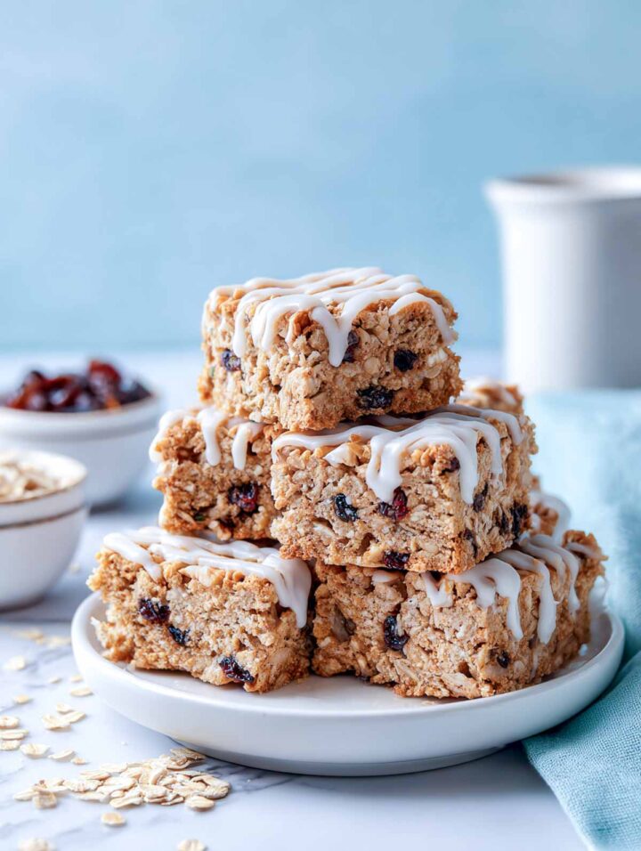 Cinnamon raisin oat squares topped with a light honey glaze, arranged on a white marble surface with a soft sage napkin. Small white bowls of oats and raisins sit nearby, creating a clean, fresh, bakery-style presentation.