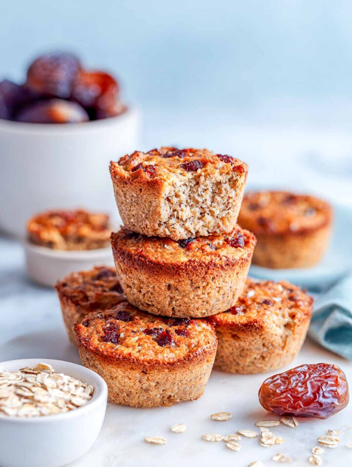 Warm date oat cakes baked in a muffin-style shape and stacked on a white marble surface. The cakes have a golden oat crumb with visible chopped dates throughout. Small white bowls of rolled oats and whole dates sit nearby with a soft sage napkin in the background, creating a bright, cozy presentation.