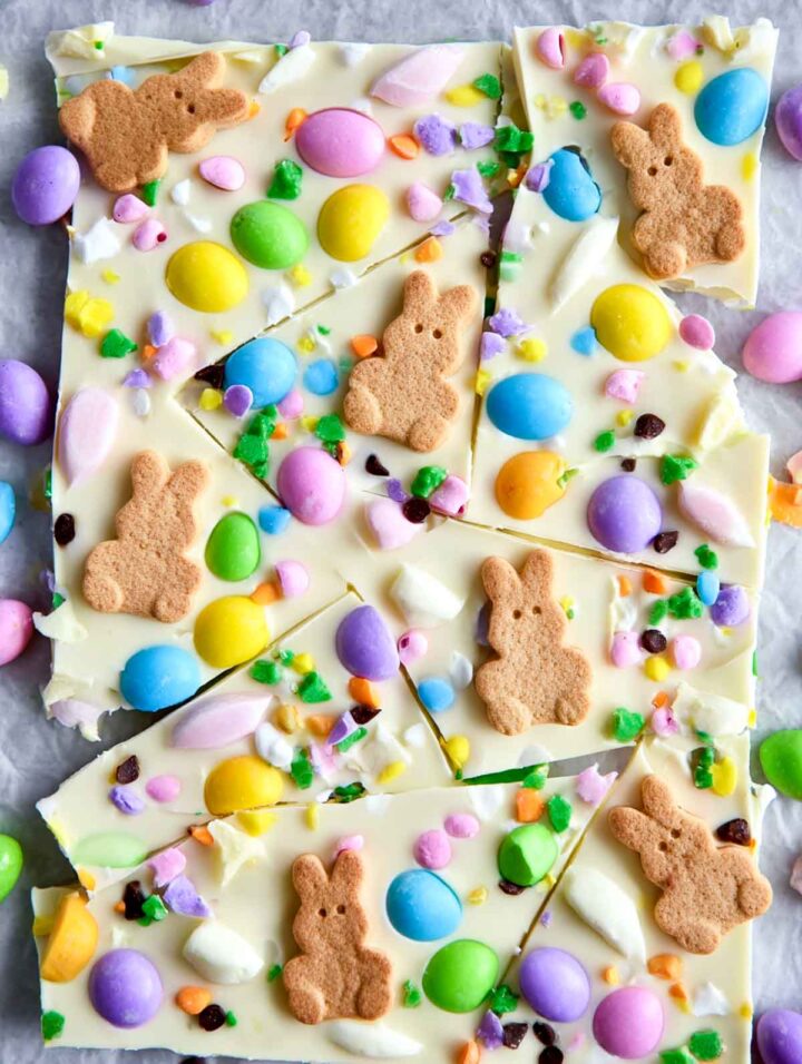 Top-down view of white chocolate Easter bark broken into pieces, topped with bunny-shaped crackers, pastel candy eggs, colorful candy pieces, and green sprinkles on parchment paper.