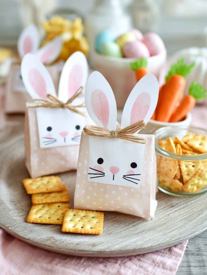 Easter bunny snack bags filled with crackers and mixed treats, decorated with paper bunny ears and faces, arranged on a light wooden board with pastel eggs and small snack bowls in the background.