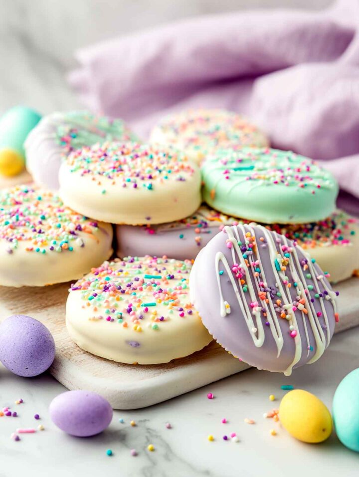 Fully dipped Oreo cookies coated in white and pastel-colored chocolate, topped with Easter sprinkles, arranged on a white marble surface with pastel mini chocolate eggs and a soft lavender napkin in the background.
