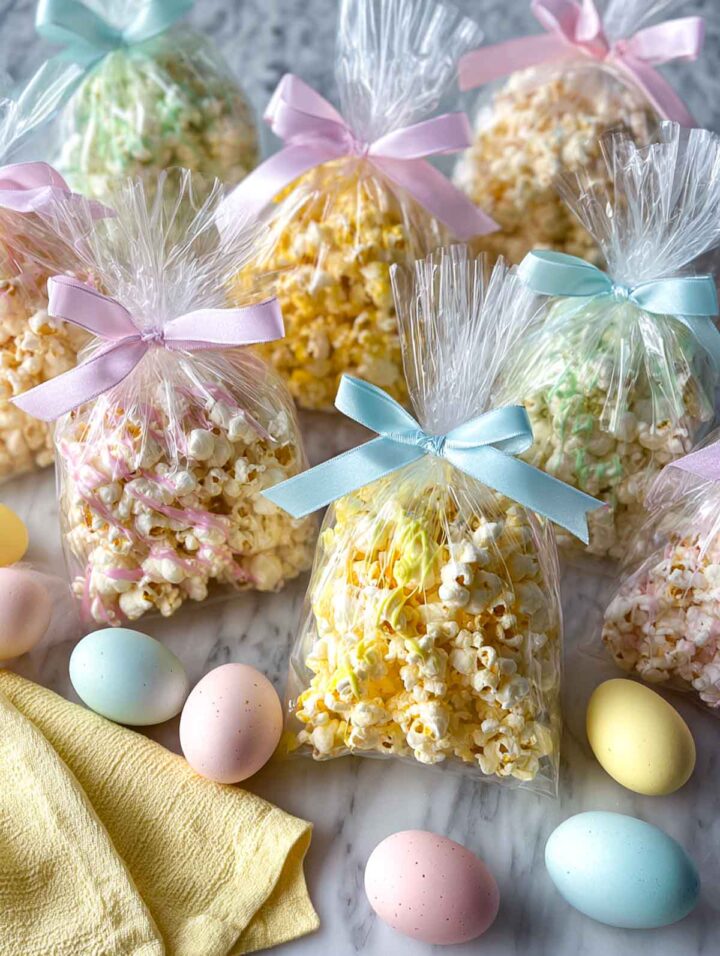 Clear cellophane bags filled with popcorn drizzled in a single pastel color, tied with soft ribbon and arranged on a white marble surface with pastel Easter eggs nearby.