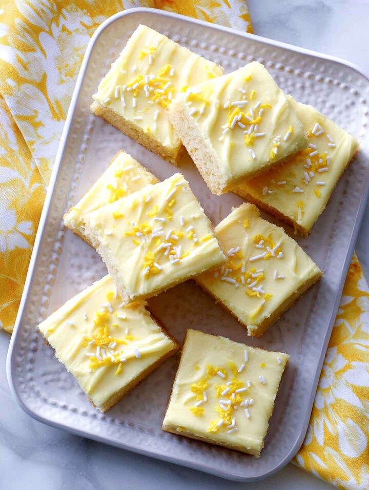 Yellow frosted sugar cookie bars arranged on a rectangular textured white serving plate, topped with yellow and white sprinkles, styled on a white marble surface with a yellow napkin and pastel Easter eggs nearby.