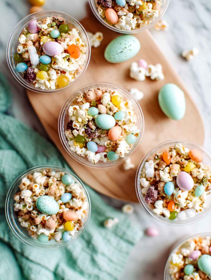 Individual clear snack cups filled with Easter trail mix made from popcorn, jelly beans, pastel candy eggs, and mixed snack pieces, arranged on a white marble surface with a light wood board, soft green napkin, and scattered pastel Easter eggs nearby.