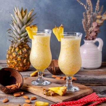 Two hurricane glasses filled with frozen piña colada, garnished with pineapple wedges, on a wooden board with a whole pineapple and almonds in the background.