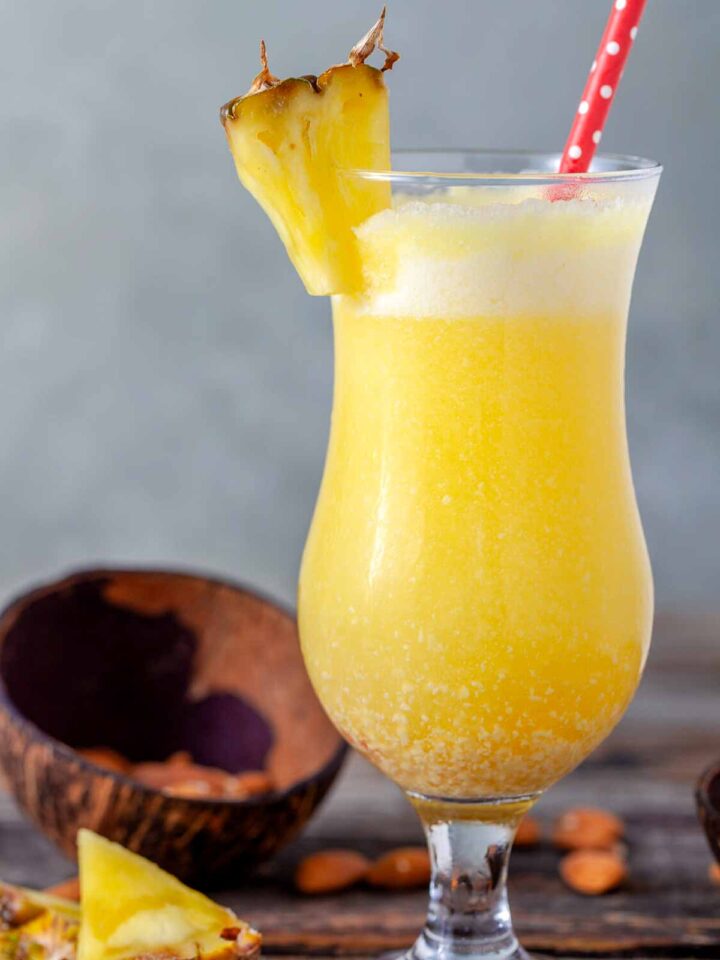Close-up of a frozen piña colada in a hurricane glass with a pineapple wedge garnish and red polka dot straw, on a wooden surface with coconut shell and almonds in the background.