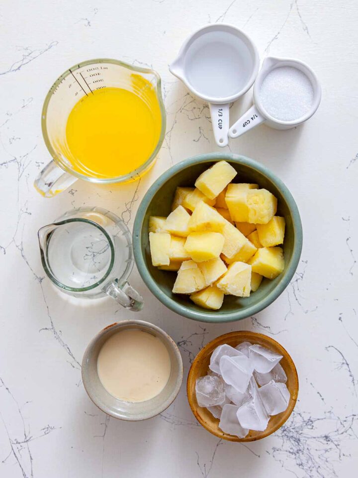 Overhead view of pineapple chunks, pineapple juice, coconut cream, water, sugar, and ice cubes arranged in bowls and measuring cups on a white marble surface.