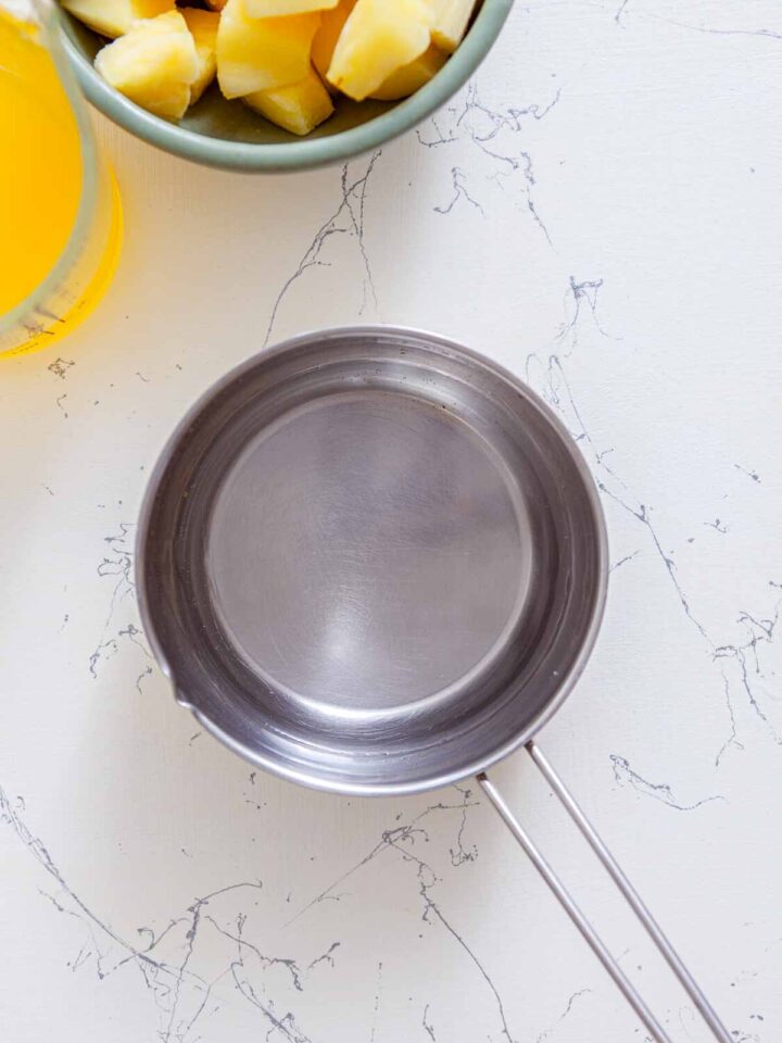Overhead view of a small stainless steel saucepan on a white marble surface with pineapple chunks and pineapple juice partially visible nearby.