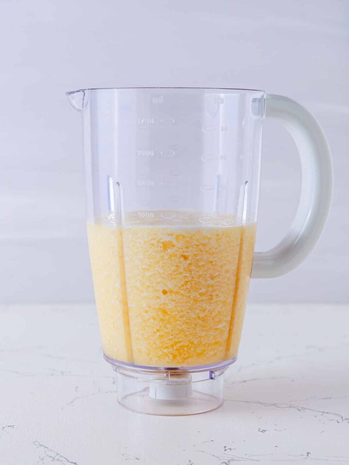 Blender pitcher filled halfway with a yellow pineapple mixture on a white marble surface.