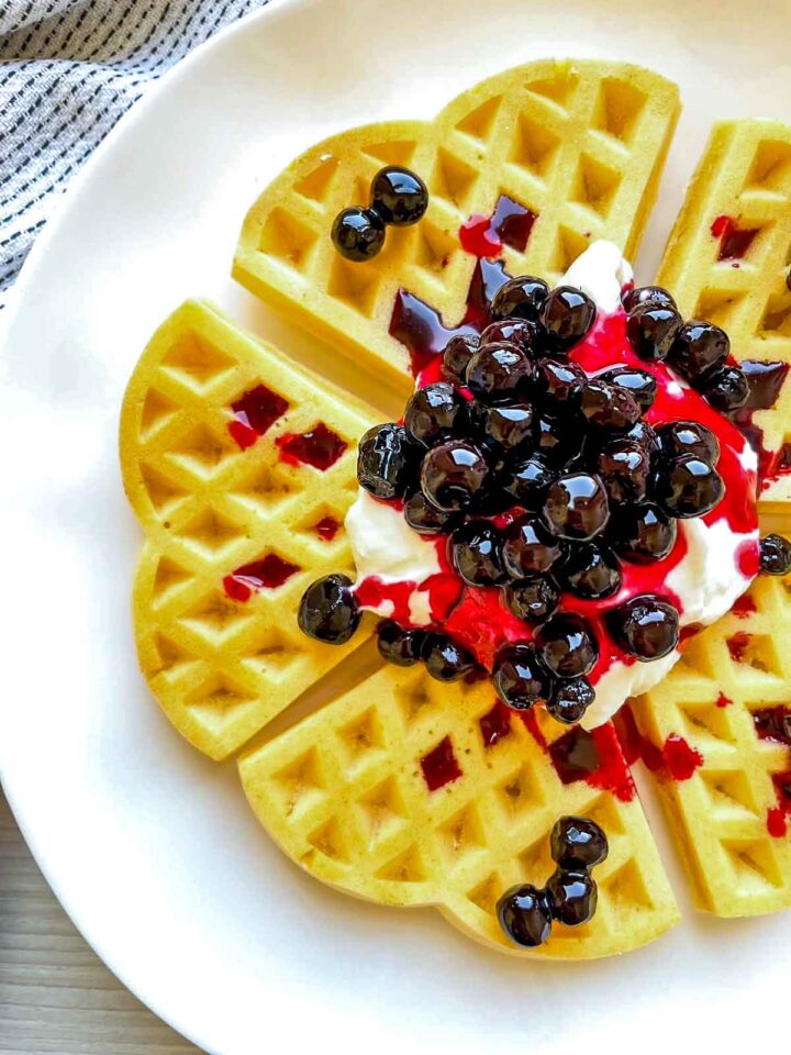 Detailed close-up of a golden waffle topped with creamy cheesecake filling and glossy blueberry sauce, highlighting the texture and vibrant colors.