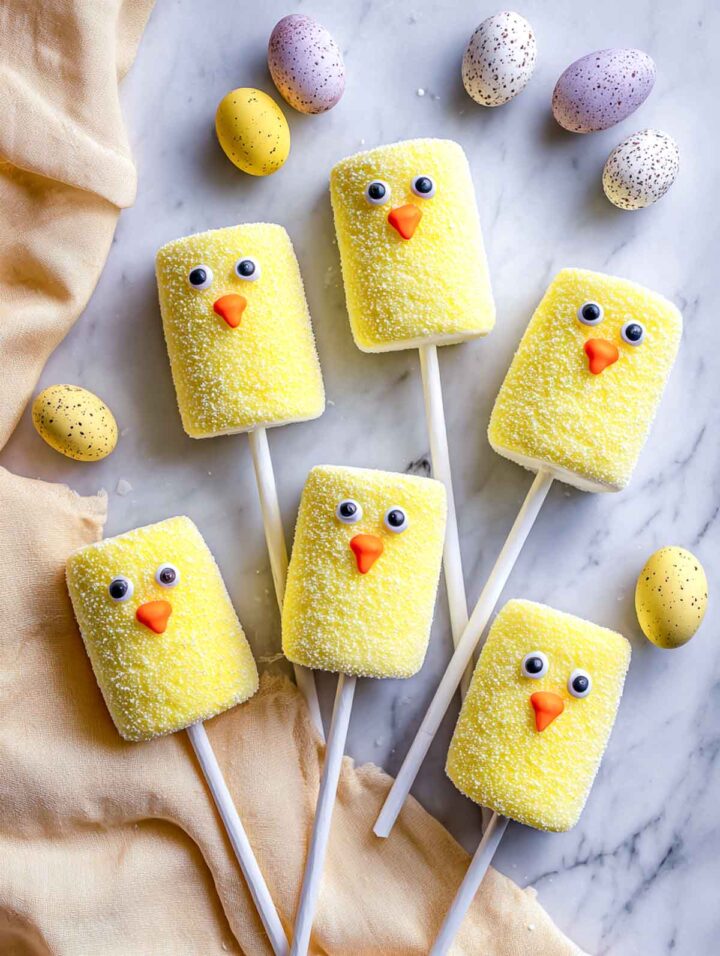 Top-down view of marshmallow Easter chick pops made from yellow sugar-coated marshmallows with candy eyes and orange beaks, attached to white lollipop sticks and arranged on a white marble surface with a soft yellow napkin and speckled pastel Easter eggs.
