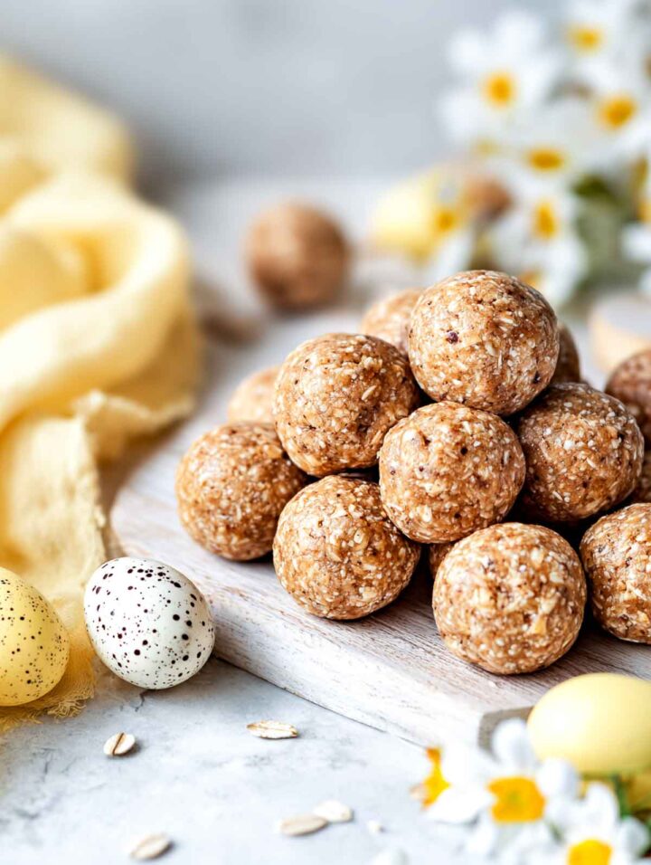 Peanut-free energy bites made with oats and honey, rolled into round balls and stacked on a light wood board on a white marble surface, with pastel Easter eggs, a soft yellow napkin, and small spring flowers in the background.
