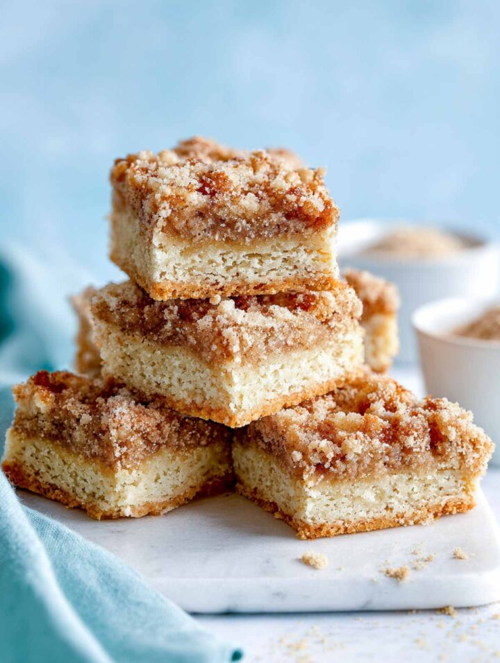 Oat bran snickerdoodle bars cut into neat squares and arranged on a white marble surface with a soft blue napkin. The bars have a pale golden crumb with visible oat bran texture and a cinnamon sugar topping that looks lightly crackled and evenly baked.