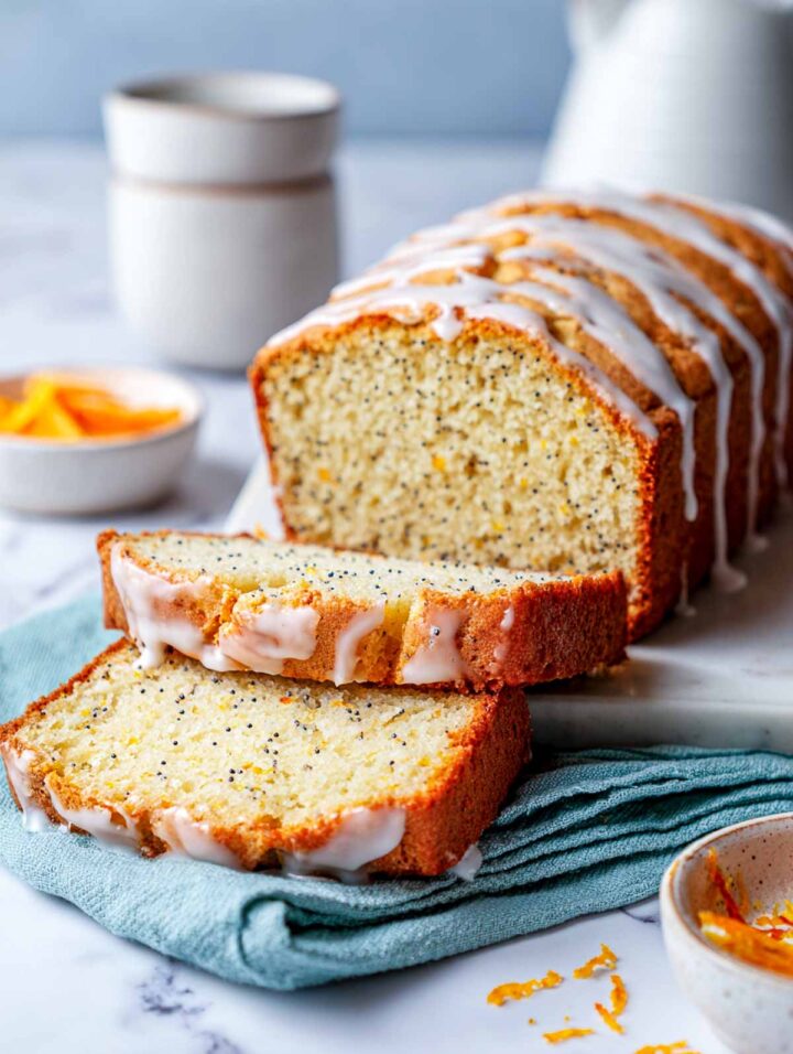 Sliced orange poppy seed yogurt loaf with a light orange glaze, shown on a white marble surface with a soft sage napkin underneath. The loaf has a pale golden crumb with evenly distributed poppy seeds and a moist texture. 