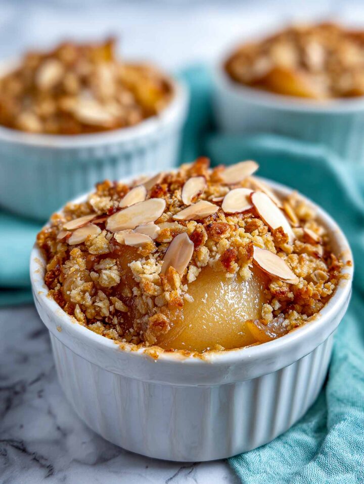 Pear almond crumble baked in a white ceramic ramekin, filled nearly to the rim with tender pear pieces and a golden oat crumble topping. Sliced almonds are visible on the surface, and the crumble looks crisp and textured. The ramekin sits on a white marble surface with a soft blue cloth in the background, creating a bright, clean presentation.