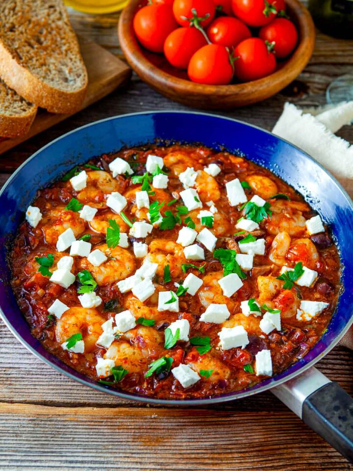 Shrimp saganaki in a skillet with tomato sauce, topped with feta cheese cubes and fresh parsley, served with bread and cherry tomatoes.