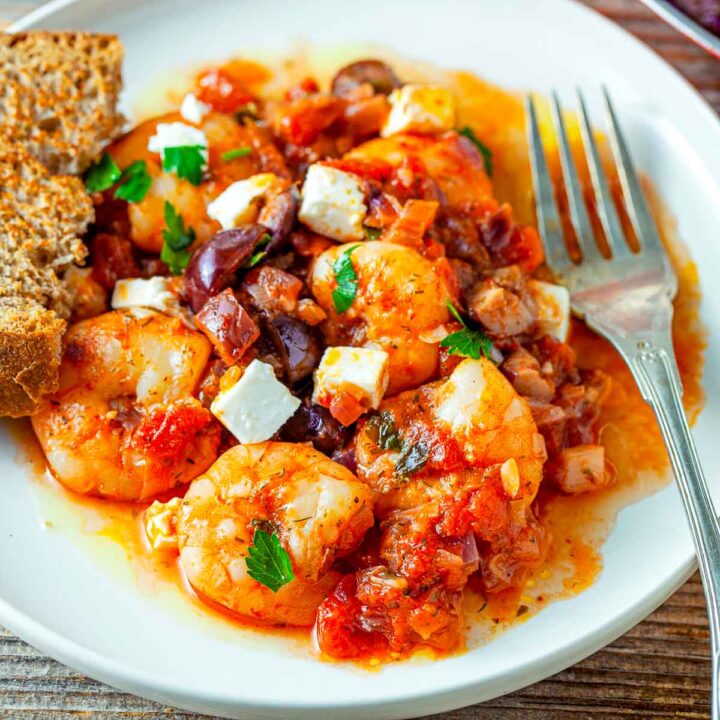 Close-up of shrimp saganaki with tomato sauce, red onions, olives, feta cubes, and parsley on a white plate, served with crusty bread and a fork.