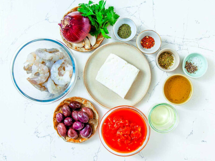 Ingredients for shrimp saganaki including raw shrimp, feta cheese, diced tomatoes, red onion, garlic, olives, olive oil, herbs, and spices arranged on a countertop.