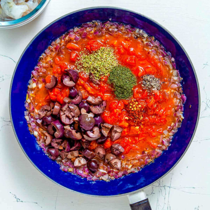 Tomatoes, olives, and herbs added to sautéed red onions in a skillet.