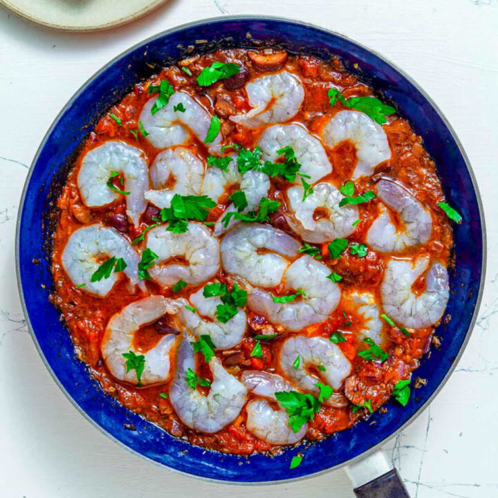 Raw shrimp arranged over tomato and olive sauce in a blue skillet, topped with fresh parsley.