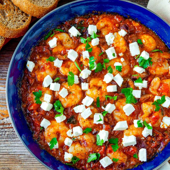 Cooked shrimp saganaki in a blue skillet, topped with feta cubes and fresh parsley, served with crusty bread.