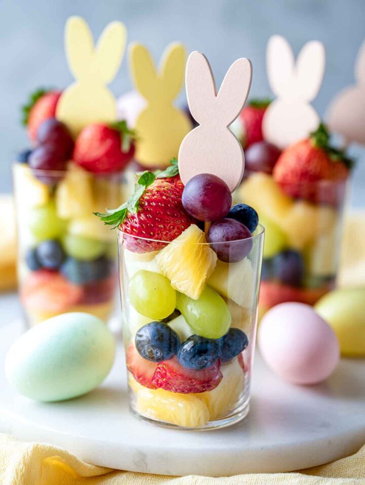 Tall, clear cups filled with layered fresh fruit, including strawberries, grapes, pineapple, blueberries, and kiwi, topped with a single pastel bunny-shaped pick. The cups are arranged on a marble surface with soft yellow napkins and pastel Easter eggs in the background, shown from a side angle with no blurring.