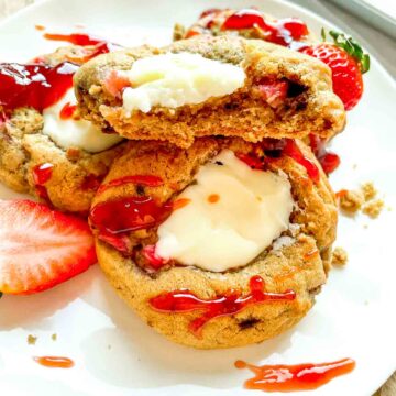 Strawberry cheesecake cookies stacked on a plate, drizzled with strawberry sauce and filled with a creamy cheesecake center