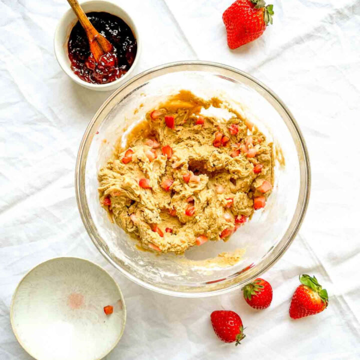 Cookie dough mixed with chopped strawberries in a glass bowl, with strawberry jam and fresh strawberries nearby