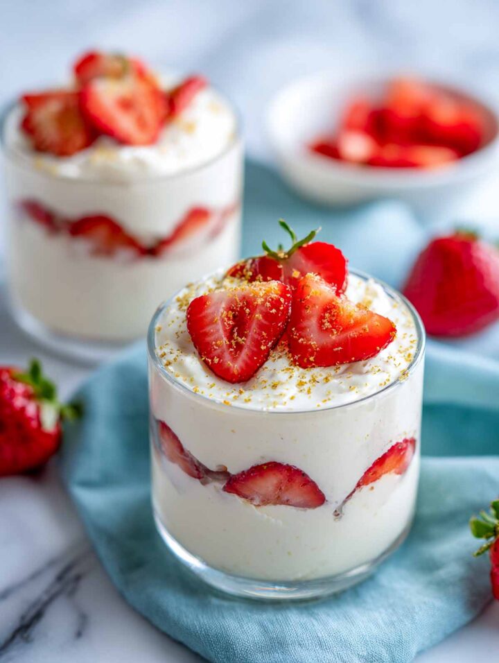 Layered strawberry ricotta cups in clear glasses with creamy ricotta filling, fresh strawberry slices pressed against the glass, and a light citrus zest topping on a marble surface.
