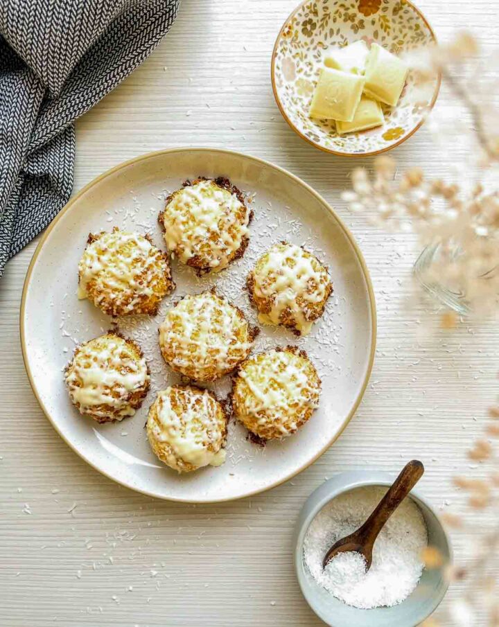 Plate of tequila white chocolate coconut macaroons topped with a white chocolate drizzle, styled with coconut flakes and white chocolate pieces