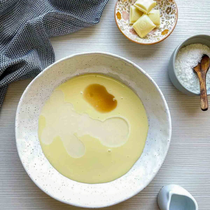 Melted white chocolate and tequila mixture in a speckled ceramic bowl, with white chocolate chunks and shredded coconut nearby on a light wooden surface