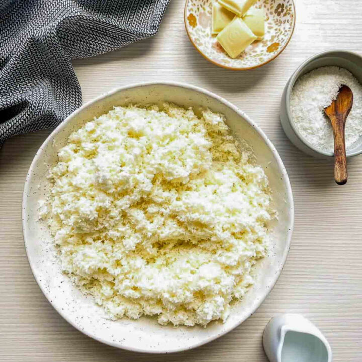 Shredded coconut mixed with white chocolate and tequila in a speckled bowl, with white chocolate pieces and coconut nearby
