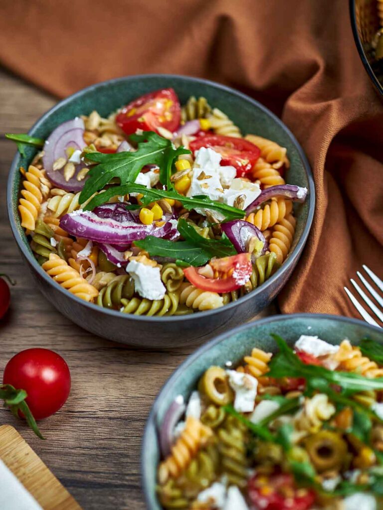 Close-up of a pasta salad in a bowl with rotini, cherry tomatoes, red onion slices, arugula, corn, seeds, and crumbled feta, set on a wooden table with a second bowl partially visible.