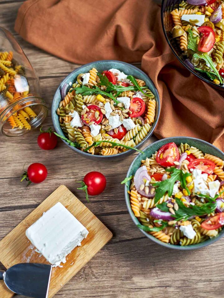 Close-up of a pasta salad in a bowl with rotini, cherry tomatoes, red onion slices, arugula, corn, seeds, and crumbled feta, set on a wooden table with a second bowl partially visible.