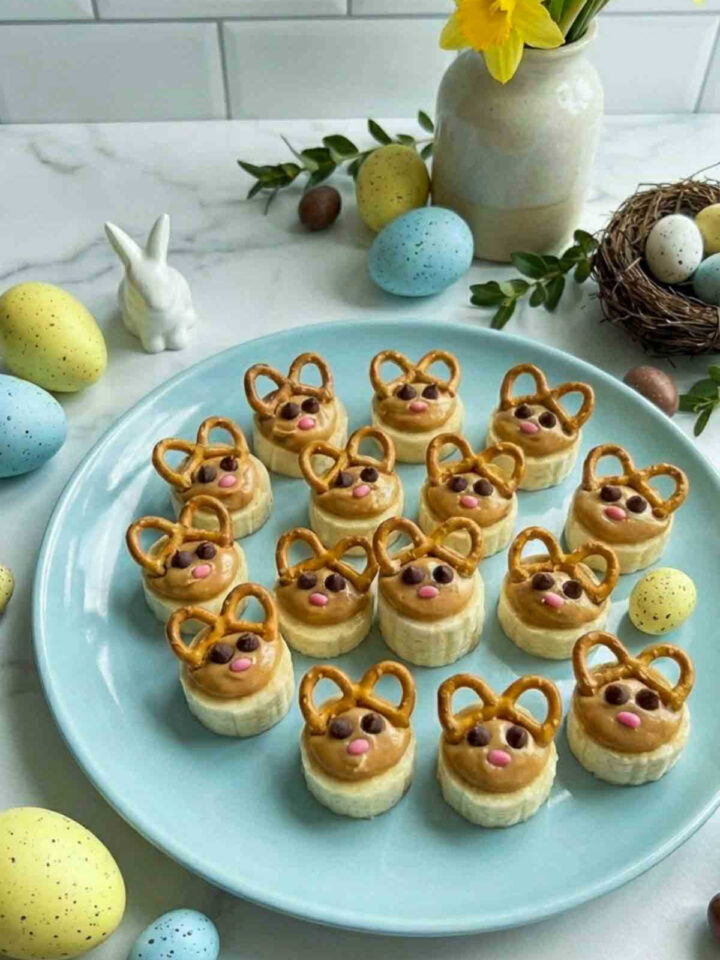 Side view of banana bunny bites arranged on a light blue plate on a white marble countertop, each banana slice topped with peanut butter, pretzel ears, chocolate chip eyes, and a small pink candy nose, surrounded by pastel Easter eggs, a small bunny figurine, and spring flowers.