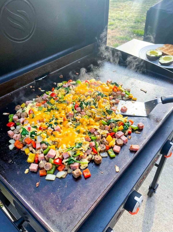 High protein breakfast scramble with eggs, sausage, ham, peppers, and melted cheese cooking on a Blackstone griddle.
