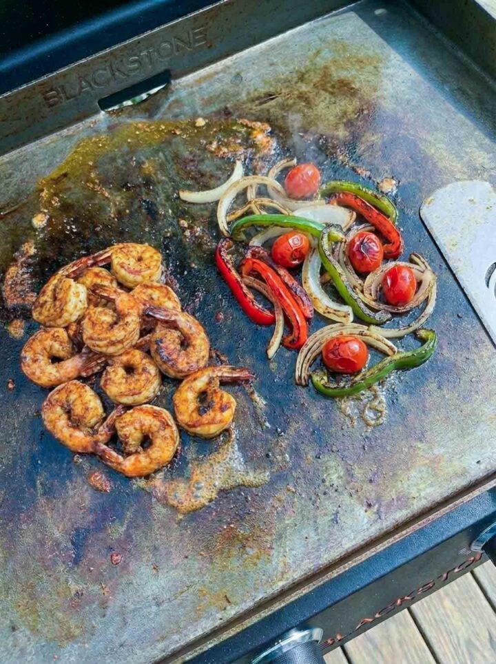 Cajun shrimp with peppers, onions, and cherry tomatoes cooking on a Blackstone griddle with light char.